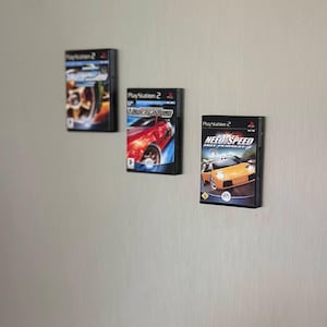 May include: Three PlayStation 2 game cases are mounted on a light gray wall. The game cases feature colorful graphics of cars and the game titles "Need for Speed Hot Pursuit 2" and "Underground".