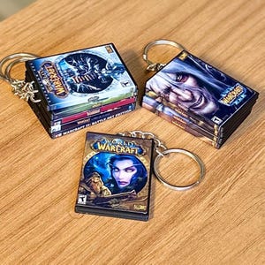 May include: Miniature World of Warcraft game box keychains. Each keychain features a stack of game boxes with different cover art. The keychains have silver-colored rings and chains. The boxes are in various colors, including blue, purple, and green.