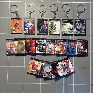 May include: A collection of miniature PlayStation 2 game case keychains. The keychains feature various game titles, including "Grand Theft Auto," "Hitman," and "Silent Hill." Each keychain has a silver keyring attached.