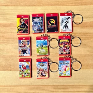 May include: A collection of red video game cartridge keychains featuring various game titles like "Cyberpunk," "Pokemon," and "Mortal Kombat." Each keychain has a silver ring. The keychains are arranged on a light brown wooden surface.