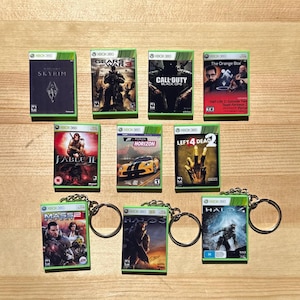 May include: Collection of Xbox 360 game keychains. The keychains feature game box art from titles like "Skyrim", "Gears of War 3", "Call of Duty: Black Ops", "Fable II", "Forza Horizon", "Left 4 Dead 2", "Mass Effect 2", "Halo 3", and "Halo 4".