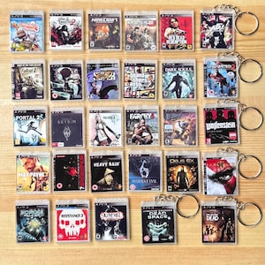 May include: Collection of PlayStation 3 game case keychains. Each keychain features a miniature game case with titles like "Minecraft", "Red Dead Redemption", and "Fallout 3". The keychains are attached to silver key rings.