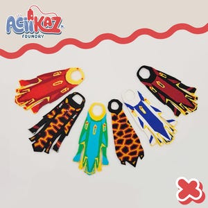 May include: A collection of colorful, felt superhero capes in red, black, blue, green, and orange. Each cape features unique geometric designs and a circular neck opening. The "AciiKaz Foundry" logo is visible in the upper left corner.