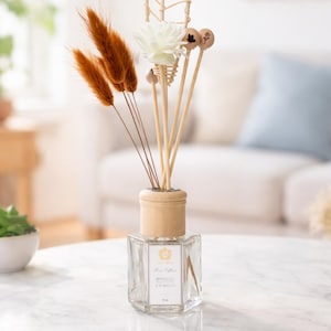 May include: A clear glass essential oil diffuser with a wooden top and decorative reeds. The diffuser is on a white marble table. The reeds include dried orange grasses, wooden sticks, and a white flower. The label reads "Home Scent".