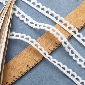 Pu&ograve; includere: Primo piano di un bordo in pizzo bianco smerlato su un righello di legno. Il pizzo ha un aspetto morbido e strutturato ed &egrave; disposto diagonalmente. Il righello mostra le misure in centimetri.