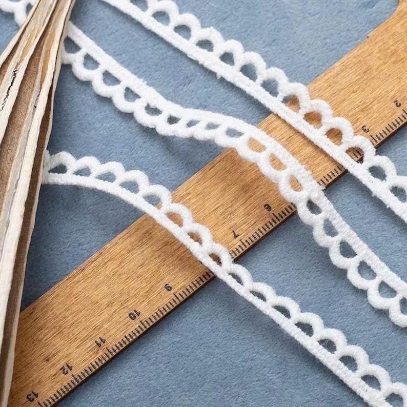 Pu&ograve; includere: Primo piano di un bordo in pizzo bianco smerlato su un righello di legno. Il pizzo ha un aspetto morbido e strutturato ed &egrave; disposto diagonalmente. Il righello mostra le misure in centimetri.