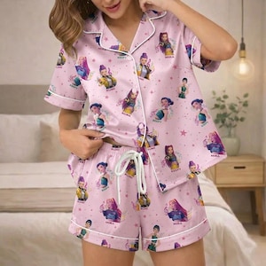 May include: Pink pajama set featuring a repeating pattern of cartoon characters, musical notes, and stars. The short-sleeved top and shorts have white piping and a drawstring waist. The set is made of a soft, silky material.