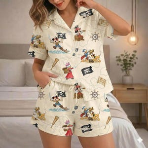 May include: Cream-colored pajama set featuring a pirate-themed print with Mickey Mouse, Minnie Mouse, Donald Duck, and Pluto. The set includes a button-down shirt and shorts, both adorned with pirate flags, treasure maps, and nautical elements.
