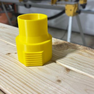 Puede incluir: Un adaptador de pl&aacute;stico amarillo brillante con una parte superior cil&iacute;ndrica y una base hexagonal con ranuras horizontales. Dise&ntilde;ado para conectar componentes, descansa sobre una superficie de madera clara.