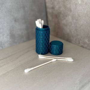 May include: A teal blue, diamond-patterned container with a matching lid, holding white cotton swabs. Three cotton swabs are placed in front of the container on a light-colored surface. The container is open, revealing the swabs inside.