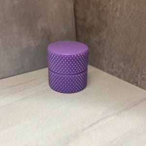 May include: A small, cylindrical container in a vibrant purple hue. The container is composed of two parts, a base and a lid, both featuring a textured, diamond-patterned surface. The object is set against a neutral background.