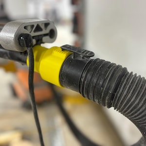 Puede incluir: Primer plano de una herramienta el&eacute;ctrica con una manguera negra corrugada conectada a un conector amarillo y negro. Un cable negro se extiende desde la herramienta. La herramienta tiene una carcasa de metal plateado.