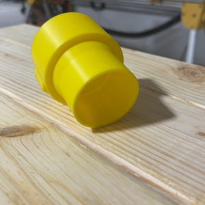 Puede incluir: Un accesorio de pl&aacute;stico amarillo brillante con forma cil&iacute;ndrica y un extremo acampanado. El accesorio descansa sobre una superficie de madera clara, probablemente un banco de trabajo. El objeto parece ser un componente de un sistema m&aacute;s grande.
