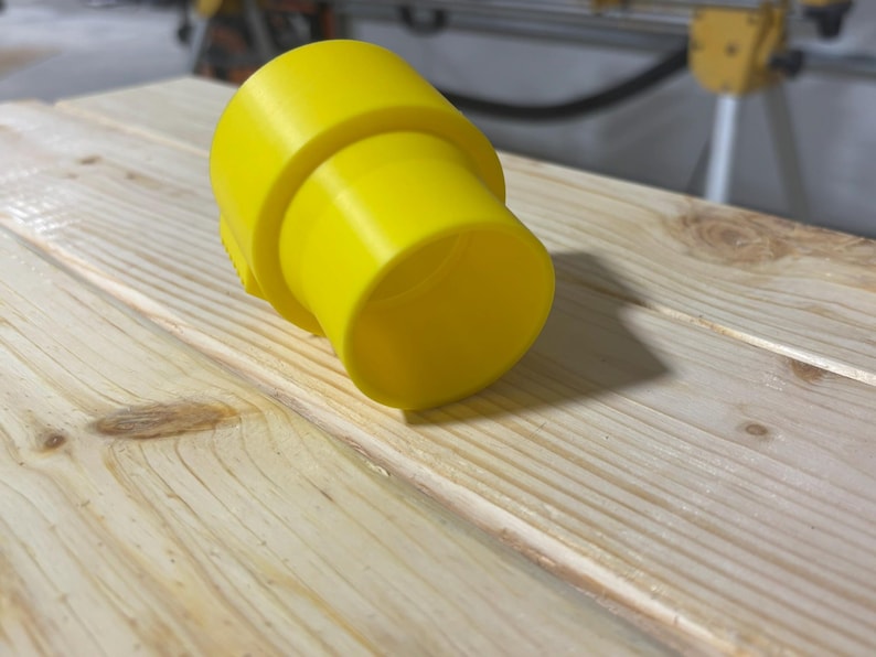 Puede incluir: Un accesorio de pl&aacute;stico amarillo brillante con forma cil&iacute;ndrica y un extremo acampanado. El accesorio descansa sobre una superficie de madera clara, probablemente un banco de trabajo. El objeto parece ser un componente de un sistema m&aacute;s grande.