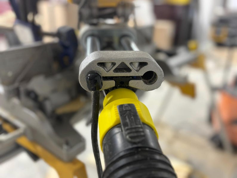 Puede incluir: Primer plano de una sierra ingletadora gris y amarilla. La sierra tiene una carcasa de metal gris con un cable de alimentaci&oacute;n negro y un conector amarillo. La sierra est&aacute; dise&ntilde;ada para cortar madera y otros materiales.