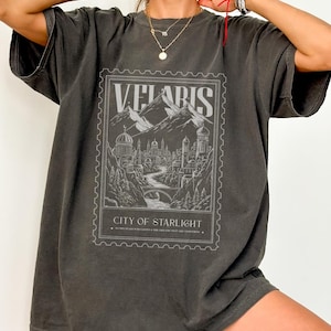 Op de afbeelding: Donkergrijs t-shirt met een gedetailleerde afbeelding van een stad tussen bergen, met de tekst "VELARIS CITY OF STARLIGHT" in een postzegelontwerp. Het shirt heeft korte mouwen en een relaxte pasvorm.