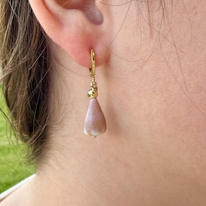 Puede incluir: Un pendiente de aro dorado con un colgante en forma de lágrima de color rosa claro. El pendiente está sujeto a la oreja. El colgante tiene una superficie texturizada y una pequeña punta blanca. El pendiente es de diseño sencillo y elegante.