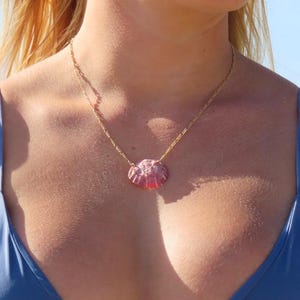 May include: A gold chain necklace featuring a pink seashell pendant. The seashell has a textured surface and is a light pink colour. The necklace is worn over a blue top.
