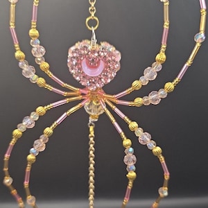 May include: A decorative spider ornament with a pink heart-shaped body and crystal accents. The spider's legs are made of gold and pink beads, and it hangs from a chain. A large crystal ball hangs below.
