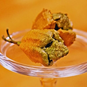 Może przedstawiać: Zbliżenie na przezroczysty szklany talerz na nóżce z kilkoma dojrzałymi, teksturowanymi paprykami. Papryki są mieszanką zieleni i pomarańczy, o pomarszczonej powierzchni. Tło jest jednolite, ciepło pomarańczowe.