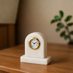 May include: A white marble mantel clock with an arched top and a rectangular base. The clock face is white with black Roman numerals and black hands, surrounded by a circular gold-toned metal bezel. The design is smooth and solid in appearance.