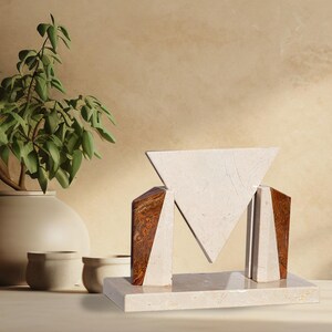 May include: A decorative sculpture featuring a central inverted triangle made of cream-colored stone, flanked by two angular supports of brown and cream stone, all mounted on a rectangular cream stone base with a smooth, polished finish.