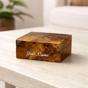 May include: A rectangular box with a polished brown and gold marbled stone finish. The lid is flat and the base features the white cursive text Your Name engraved on the front side. The box has a smooth, glossy surface with natural veining.