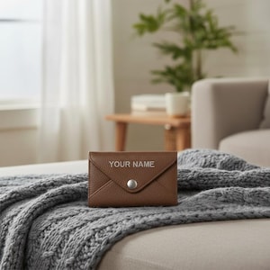 May include: A brown leather envelope-style wallet with a textured finish, featuring a silver-toned metal snap button closure on the front flap and the text "YOUR NAME" embossed in a clean, sans-serif font centered above the snap closure.
