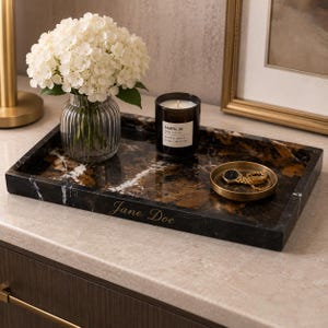 May include: A rectangular black and brown marble tray with white veining, featuring the engraved name Jane Doe on the front edge. The tray holds a clear ribbed glass vase with white hydrangea flowers, a black candle, and a small gold dish with a watch.