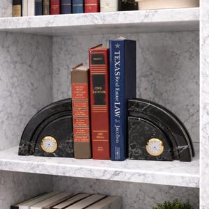 May include: Two semi-circular black marble bookends with gold-rimmed analog clocks embedded in the center. The bookends support three hardcover books, including a blue volume titled Texas Real Estate Law and a red volume titled Civil Procedure.