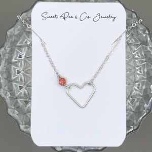 May include: A silver-toned necklace featuring a delicate chain, an open heart-shaped pendant, and a single small round orange gemstone accent. The jewelry is displayed on a white card with the text Sweet Pea & Co. Jewelry printed at the top.