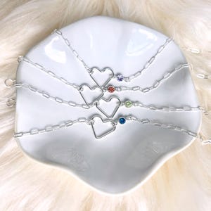 May include: Four silver-toned chain bracelets, each featuring a central open heart charm and a single small round gemstone. The gemstones are purple, orange, light green, and blue. The bracelets have a delicate link chain design with a circular clasp.