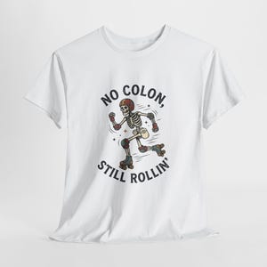 May include: A white short-sleeved t-shirt featuring a graphic of a skeleton wearing a helmet, elbow pads, knee pads, and roller skates. The text "NO COLON, STILL ROLLIN'" is printed in black capital letters in an arc around the skeleton illustration.
