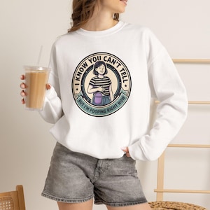May include: A white crewneck sweatshirt featuring a circular graphic with a cartoon illustration of a person in a striped shirt. The text reads I KNOW YOU CAN'T TELL BUT I'M POOPING RIGHT NOW in black lettering around the central circular design.