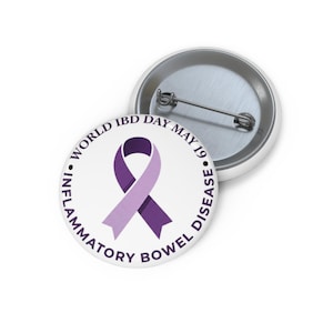 May include: A white button pin with a purple ribbon design, and the text "WORLD IBD DAY MAY 19" and "INFLAMMATORY BOWEL DISEASE" in a circular pattern. The pin has a metal back and a safety pin clasp.