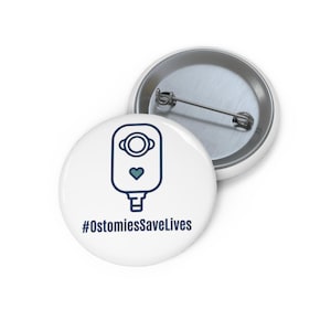 May include: Two white round buttons. One button has a blue and white graphic of a medical device with a heart and the text "#OstomiesSaveLives". The other button is silver.