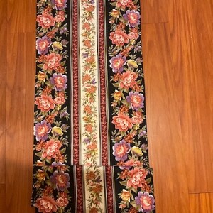 Puede incluir: Tela negra con un patrón repetitivo de flores rosas, moradas y verdes. La tela tiene tres rayas verticales blancas, rojas y crema.