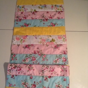 Könnte beinhalten: Ein gestreifter Tischläufer mit einem floralen Muster in Rosa-, Gelb- und Blautönen. Der Läufer ist aus Stoff und hat eine gesteppte Textur.