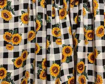Könnte beinhalten: Gardinenpaneele mit Sonnenblumenmuster. Die Gardinen zeigen ein schwarz-weißes Karomuster mit leuchtend gelben Sonnenblumen und grünen Blättern. Der Stoff ist oben gerafft, wodurch weiche Falten entstehen.