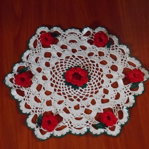 Könnte beinhalten: Weiße Häkeldeckchen mit grünem Rand und sechs roten Häkelrosen.