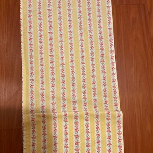 Puede incluir: Tela amarilla con finas rayas rojas y blancas y un pequeño estampado floral. La tela está doblada por la mitad.