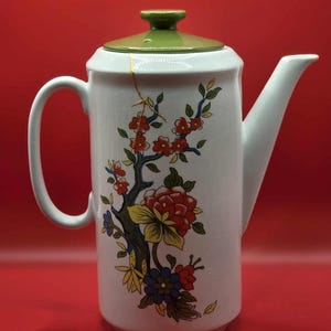 May include: White ceramic teapot with a green lid and handle, featuring a floral design in red, yellow, and blue. The teapot has a long spout and a gold repair line. The background is a solid red.