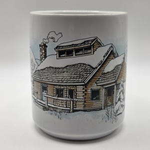 May include: White ceramic mug featuring a detailed illustration of a snow-covered cabin with a chimney and smoke. The cabin is brown with a snow-covered roof and windows. The background has a light blue and white winter scene.