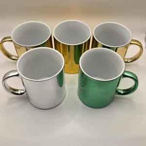 May include: Five metallic mugs in gold, silver, and green. Each mug has a white interior and a matching metallic handle. The mugs are arranged in a semi-circle, showcasing their shiny exteriors.