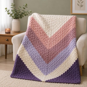 May include: A crochet blanket featuring a chevron pattern with alternating stripes of cream, dusty rose, mauve, light purple, and deep purple. The textile has a textured, waffle-like stitch throughout, creating a soft and thick appearance.