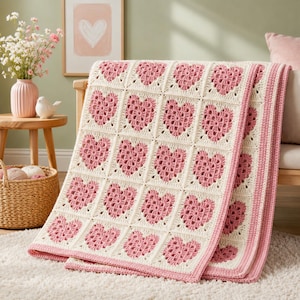 May include: A handmade crochet blanket featuring a repeating pattern of pink heart motifs centered within off-white square blocks. The blanket is finished with a solid pink border and is constructed from yarn, showcasing a textured, grid-like design.
