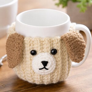 May include: A white ceramic mug featuring a removable, cream-colored crocheted cozy designed to look like a puppy. The cozy includes two brown crocheted ears, a white muzzle, a black stitched nose and mouth, and two round black plastic eyes.