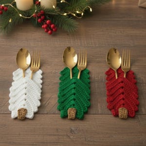 Może przedstawiać: Trzy szydełkowe uchwyty na sztućce w kształcie choinki w kolorze białym, zielonym i czerwonym, każdy z złotą łyżką i widelcem. Uchwyty mają brązową podstawę i są ułożone na drewnianej powierzchni, z świątecznymi dekoracjami w tle.