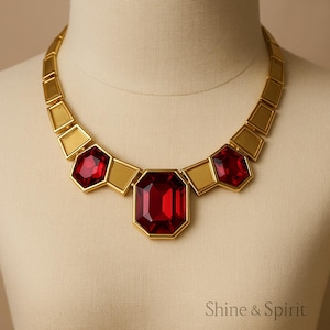 May include: A gold-toned necklace featuring large, faceted red gemstones. The necklace has a geometric design with rectangular and hexagonal shapes. The central gemstone is rectangular, flanked by two hexagonal stones, all set in gold. The necklace is displayed on a mannequin.
