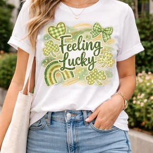 Puede incluir: Camiseta blanca con un diseño del Día de San Patricio. La camiseta presenta el texto "Feeling Lucky" en verde, junto con tréboles verdes, un arcoíris y un lazo. El diseño tiene un efecto de acuarela.